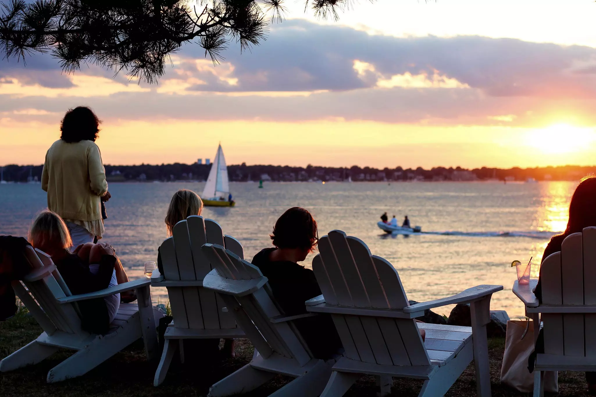 People in Adirondack chairs looking at the sunset over the Narragansett Bay in Rhode Island; a sailboat and small motorboat are in the water.