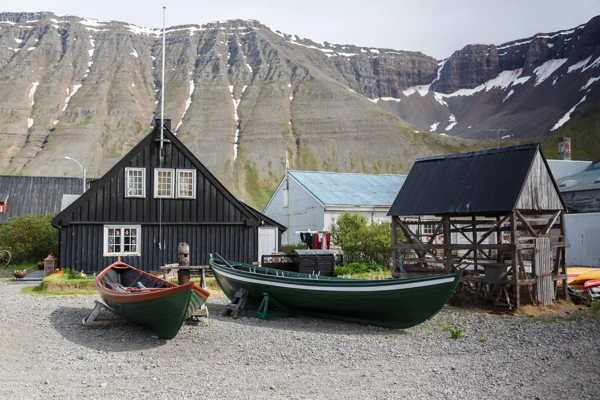 Small boats sit on a rocky beach in front of historic timber houses in Ísafjörður, Iceland. Snow-dusted cliffs rise in the background.