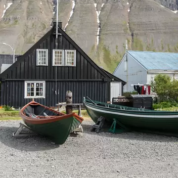 Small boats sit on a rocky beach in front of historic timber houses in Ísafjörður, Iceland. Snow-dusted cliffs rise in the background.