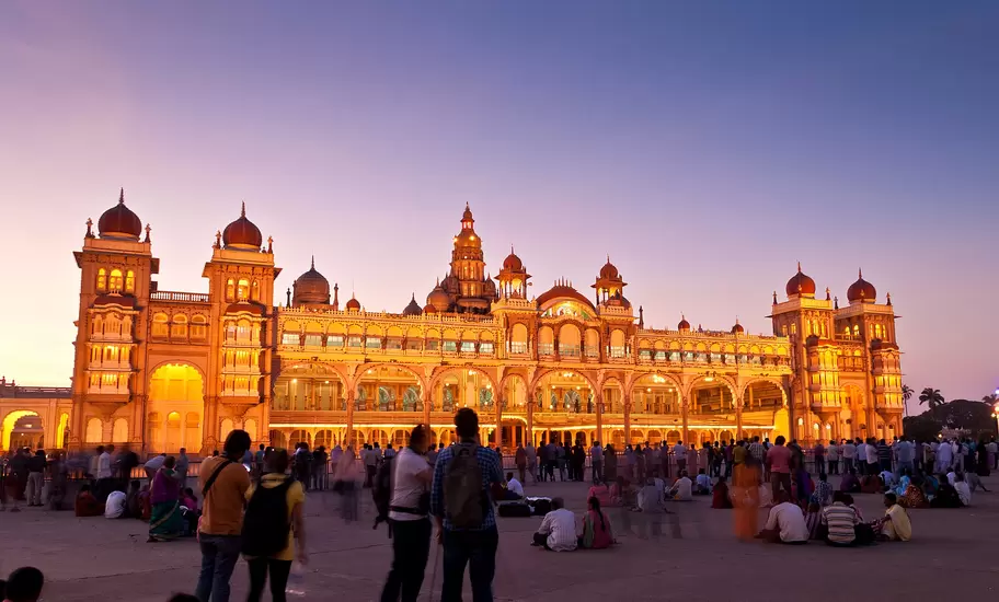 Mysuru Palace lit up in gold at dusk, its domed towers and arched facade above crowds gathered in the forecourt