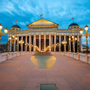 Colonnaded Archaeological Museum in Skopje at dusk, lettered in Cyrillic, reached by a bridge lined with statues and lamps