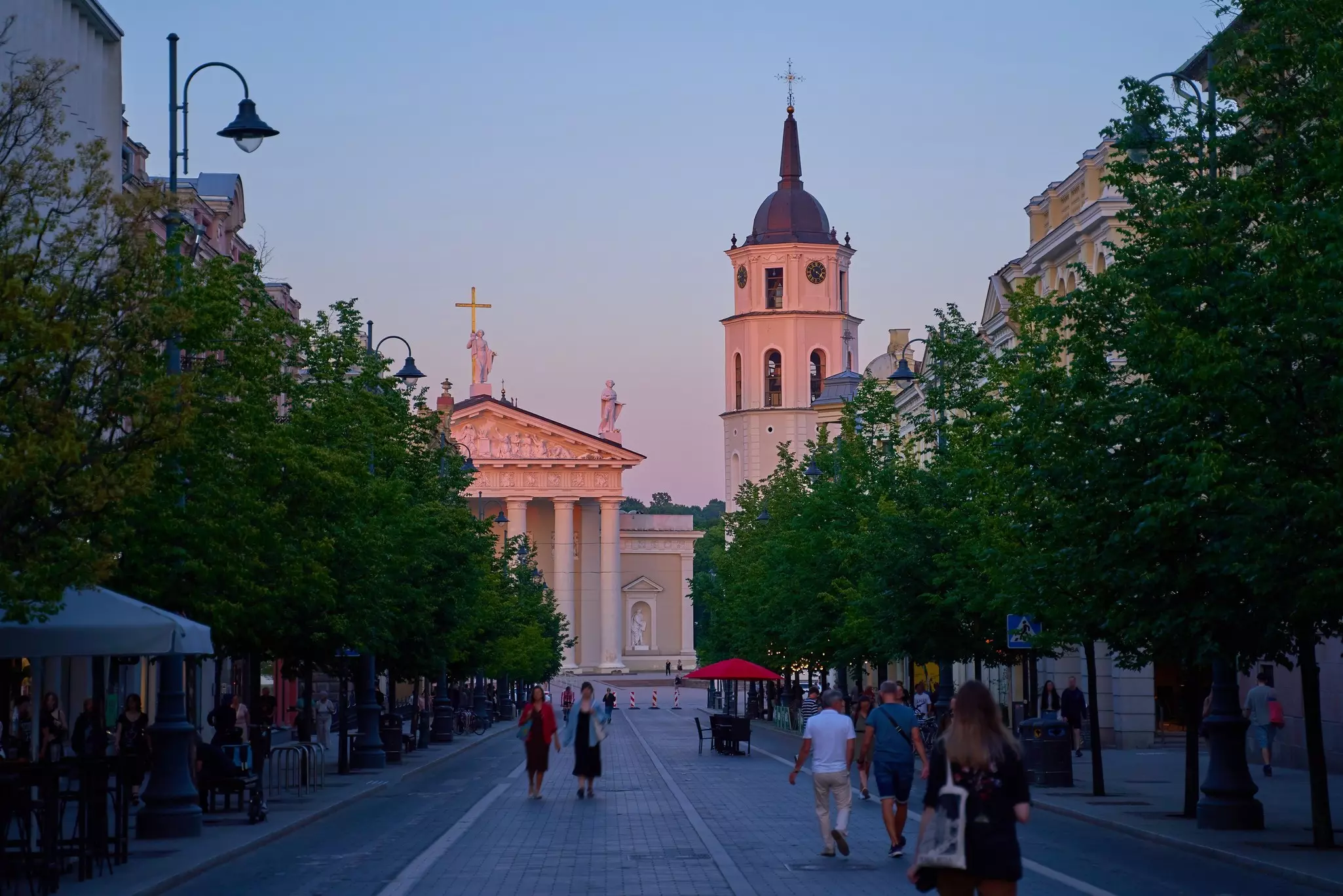 People in dark shadow walking on a broad pathway that's lined with leafy green trees, with the Vilnius Cathedral in the background in the pink light of sunset.