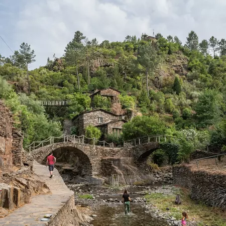 A person walking toward two stone bridges that cross a shallow stream; there are a few buildings on a hillside and two people in the stream.