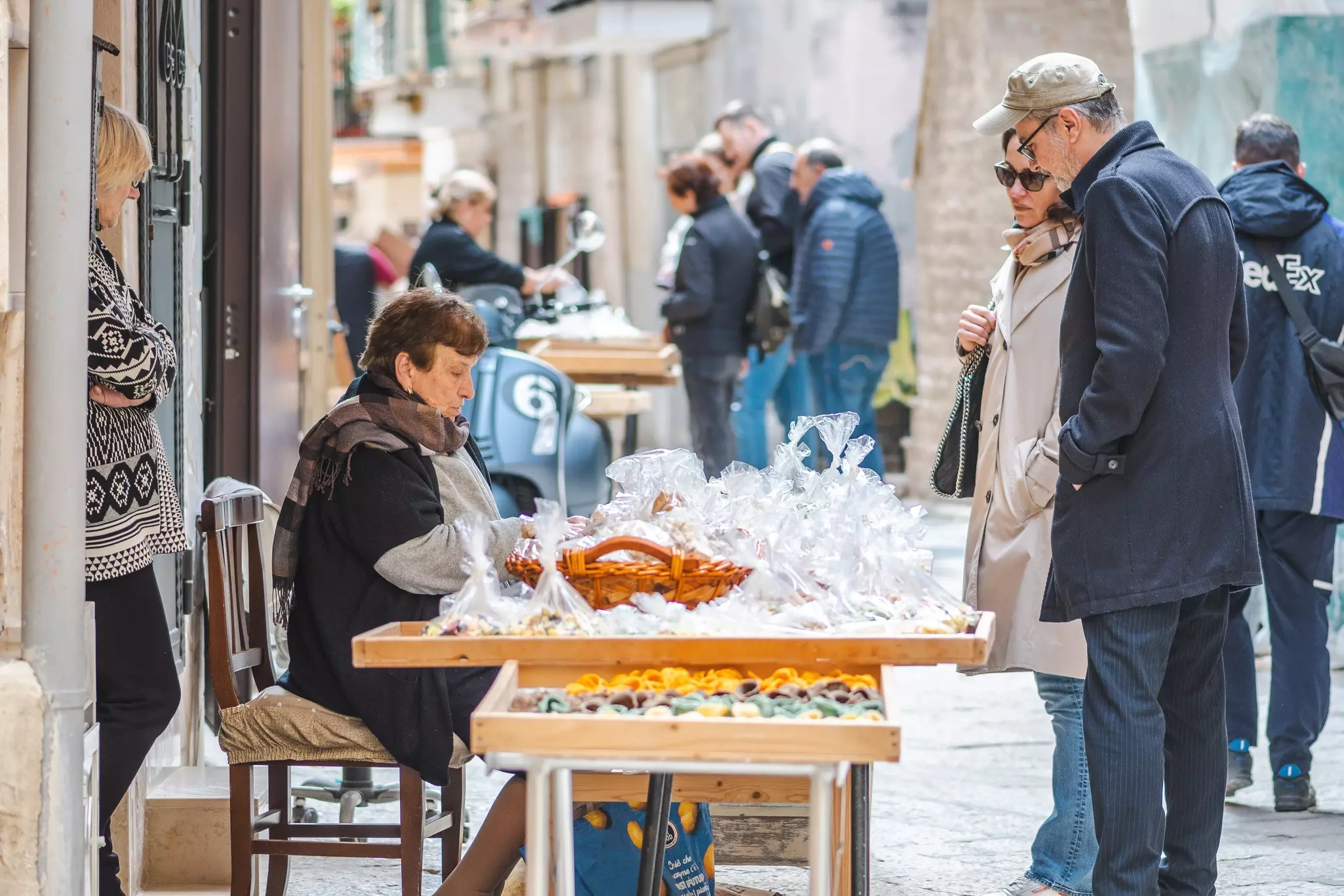 A woman wearing a scarf sits at a table set on the street with trays of fresh pasta; two customers are looking at the selection.