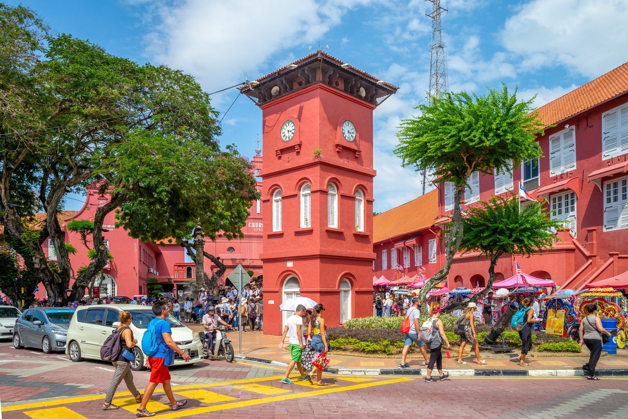 People crossing a street in front of a red colonial clock tower and matching buildings, with trees and decorated trishaws