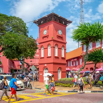 People crossing a street in front of a red colonial clock tower and matching buildings, with trees and decorated trishaws