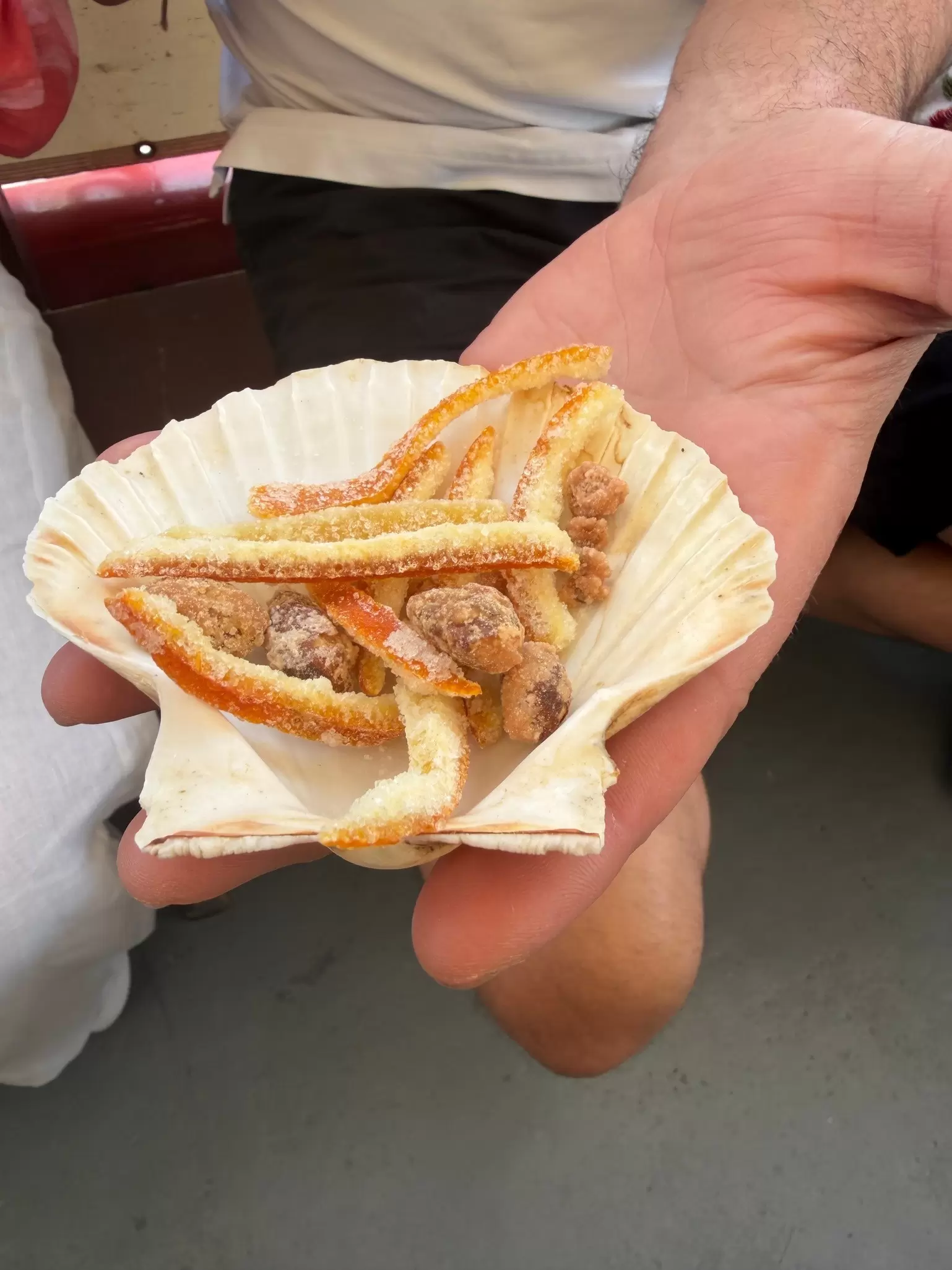 A man holds a small seashell filled with sugar orange peels and sugared almonds.