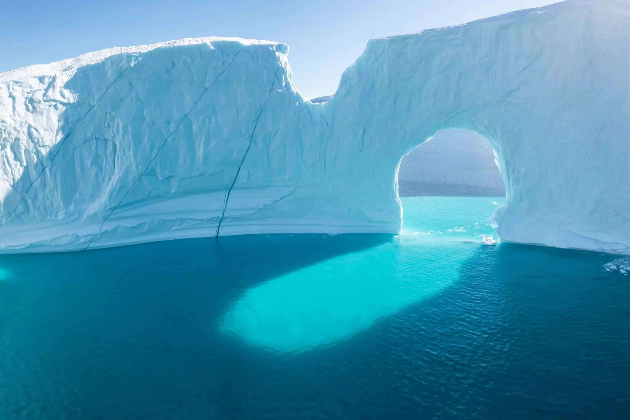 The ocean glows bright blue in the sunlight as it shines through an archway within an iceberg.