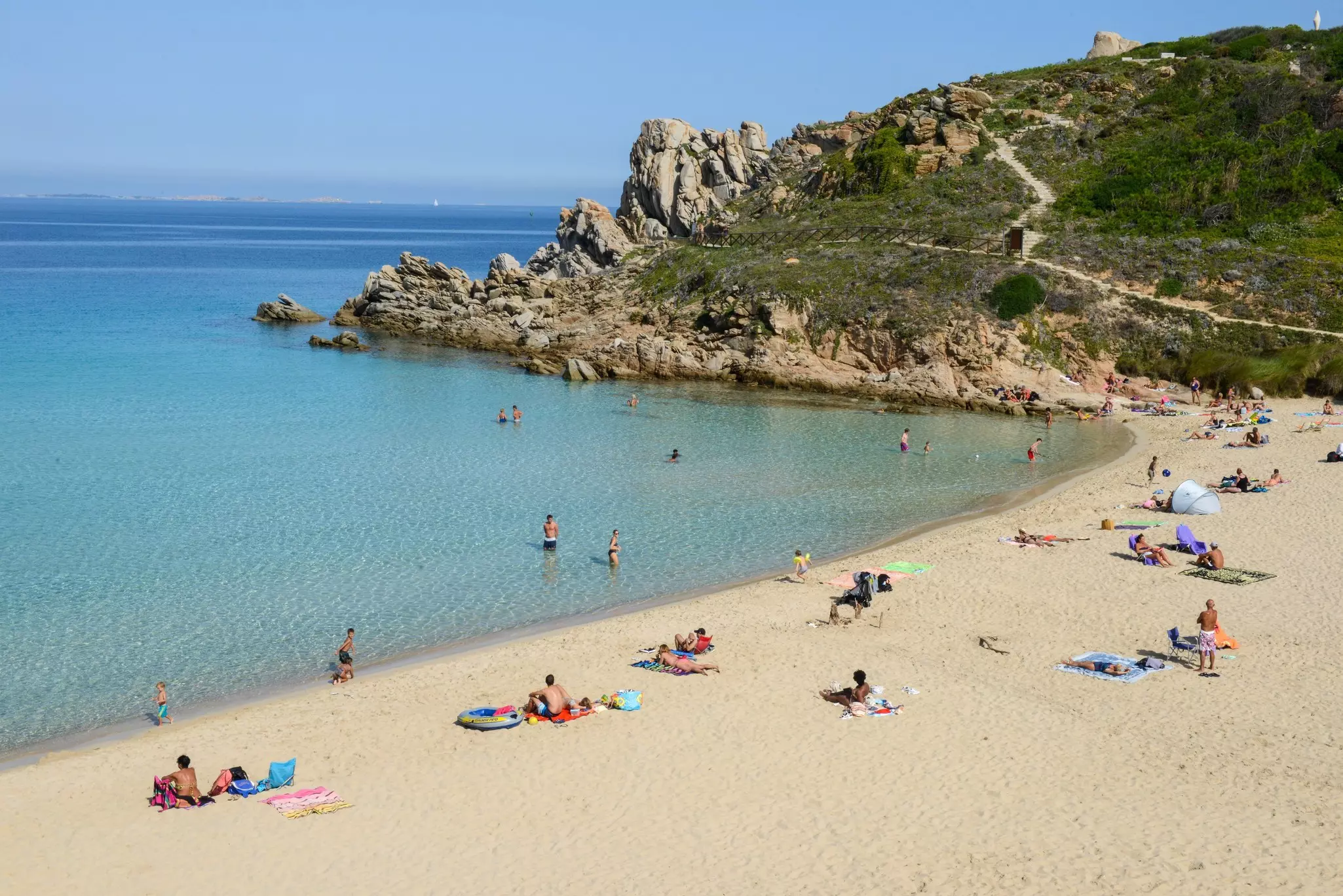 People on a white sand beach and in the clear blue water in a cove bordered by a rocky hillside.