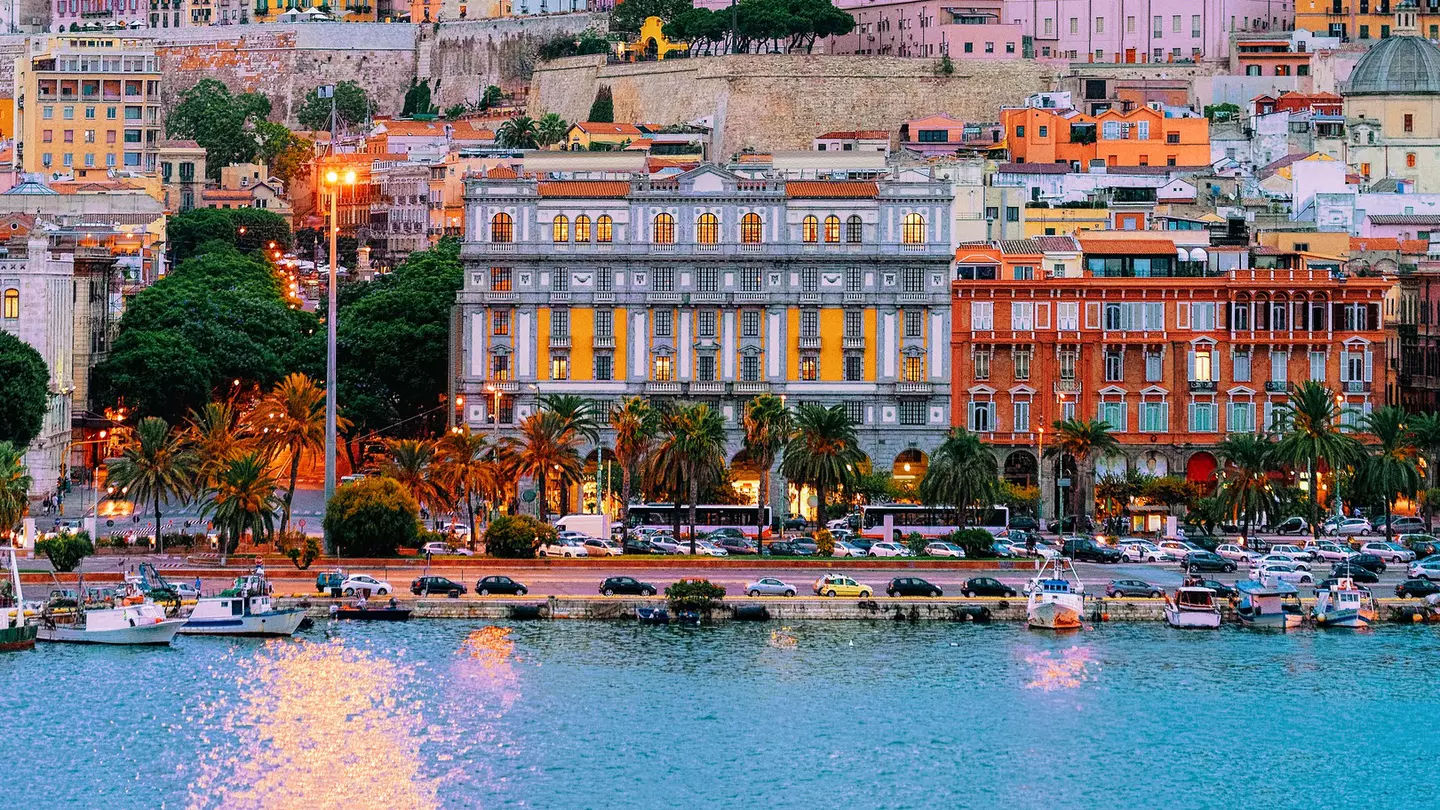 Colorful facades of buildings rising on a hill in a city. Boats are in the marina, and a road leads along the waterfront.