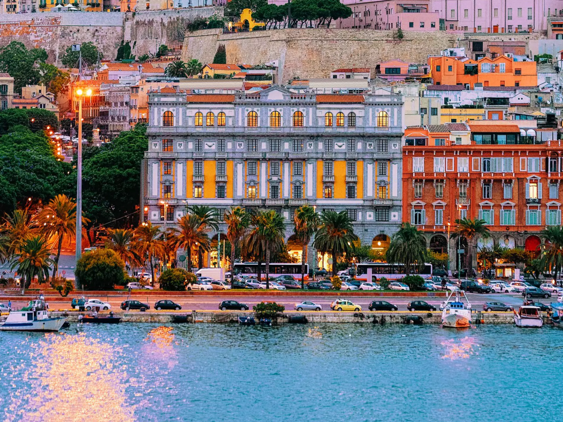Colorful facades of buildings rising on a hill in a city. Boats are in the marina, and a road leads along the waterfront.