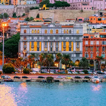Colorful facades of buildings rising on a hill in a city. Boats are in the marina, and a road leads along the waterfront.