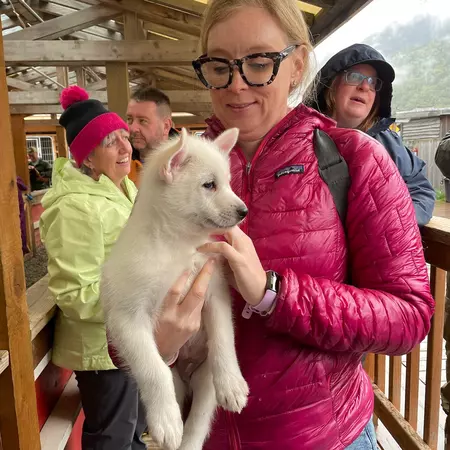 A woman wearing a red winter jacket and glasses holds a white, furry puppy.