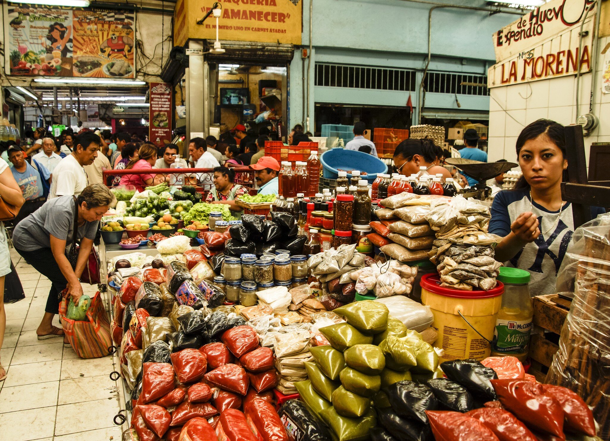 Market stall stacked with bagged spices and chillies, with shoppers and vendors in a busy covered hall.