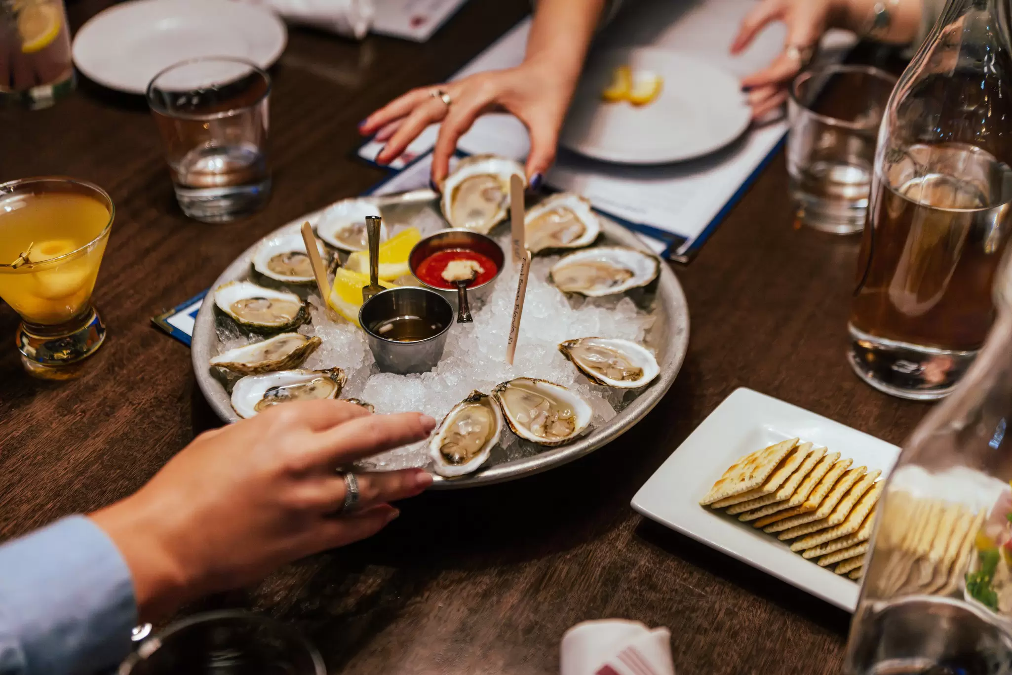 Hands reaching for oysters arranged on ice with sauces at the center 