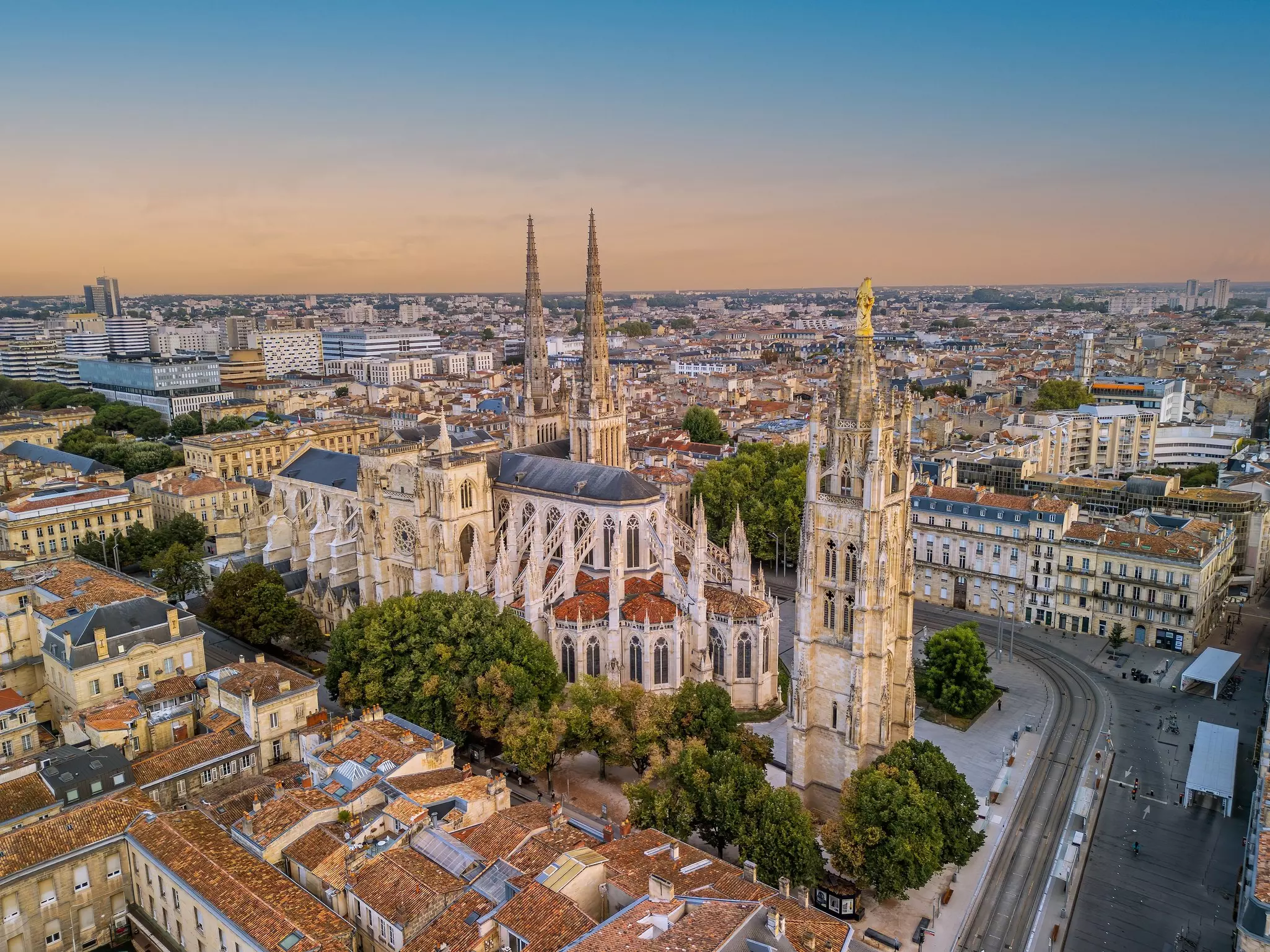 A grand cathedral with flying buttresses stands near a tall Gothic tower in a city square.