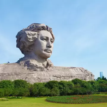 Colossal stone bust of a young Mao Zedong with wind-swept hair above trees and a flowerbed, Orange Isle, Changsha.