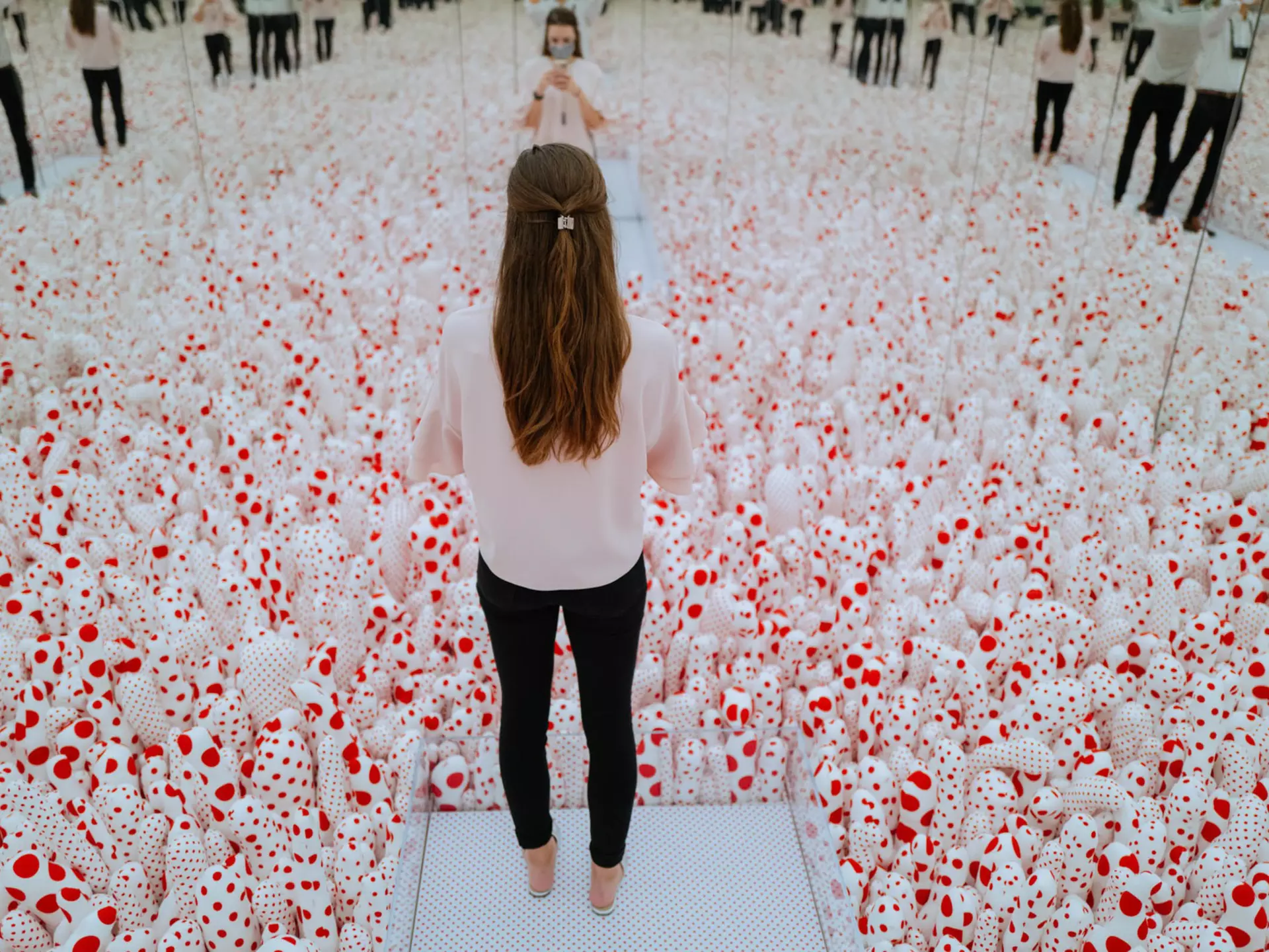 A visitor standing in Yayoi Kusama’s Infinity Mirror Room—Phalli’s Field (1965/2017), a mirror-lined room filled with phallic red-and-white polka-dotted sculptures