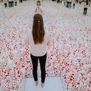 A visitor standing in Yayoi Kusama’s Infinity Mirror Room—Phalli’s Field (1965/2017), a mirror-lined room filled with phallic red-and-white polka-dotted sculptures