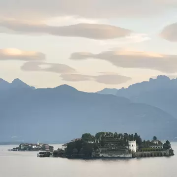 Terraced gardens and palace of Isola Bella on calm Lake Maggiore, a smaller island village to the left, hazy mountains behind.