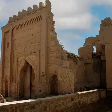 Crumbling earthen gate with a horseshoe arch, carved panels and crenellated top beside a second broken gate near Rissani