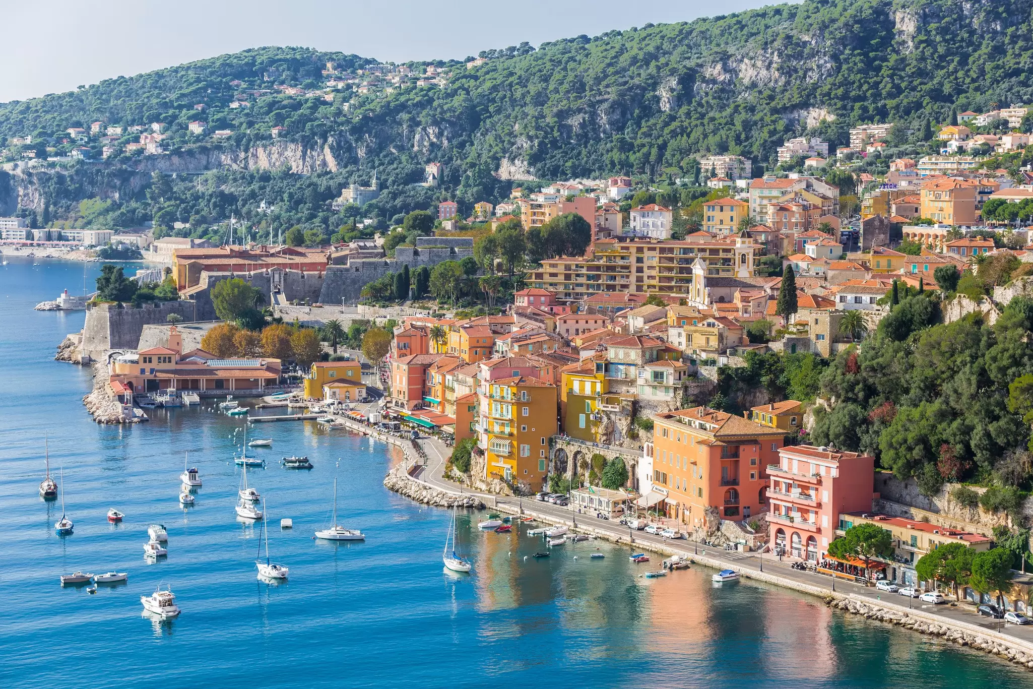 Colorful buildings on a coastline, with blue water in the foreground and green hills behind.