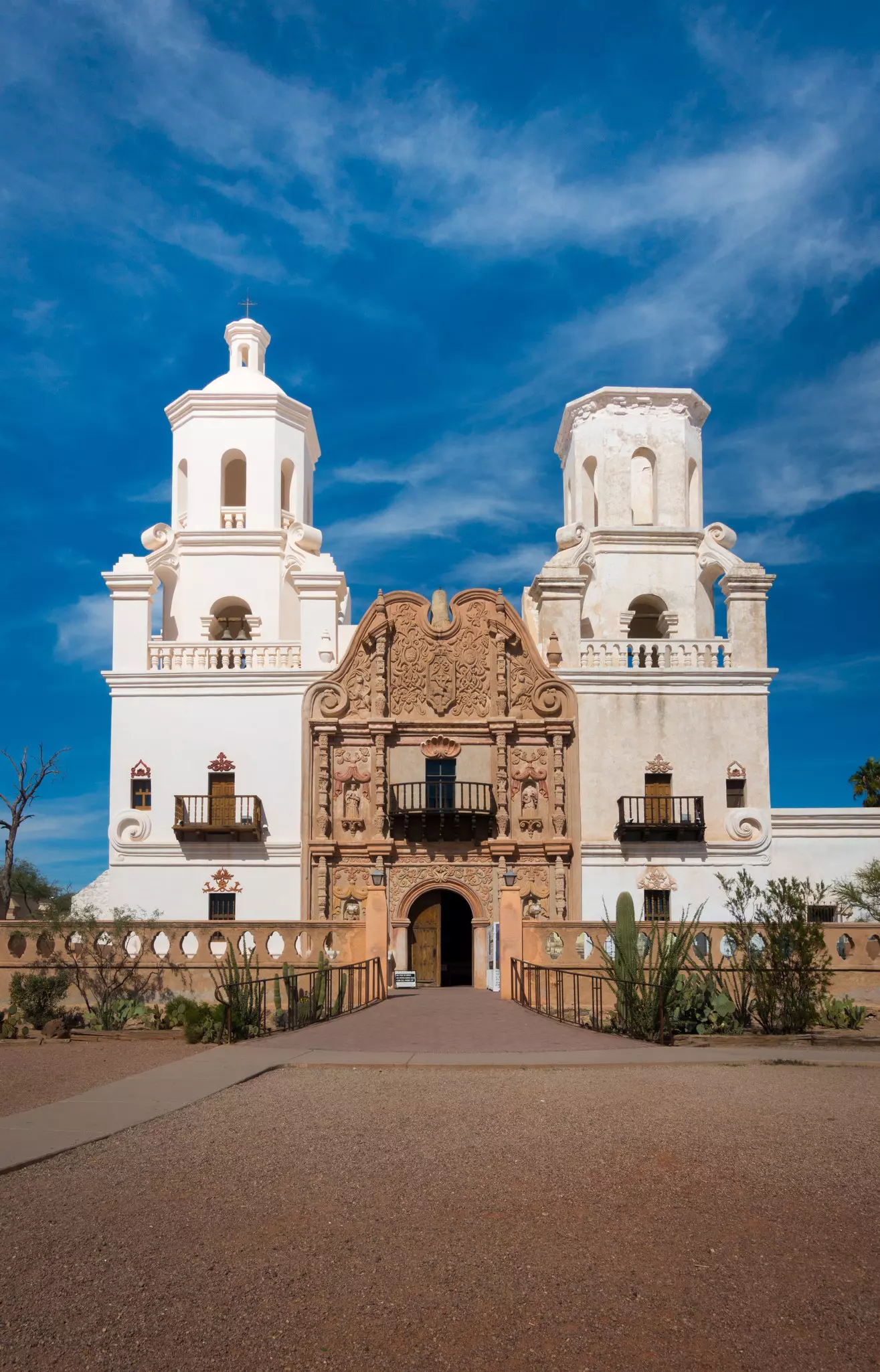 Frontal shot of Mission of San Xavier del Bac in Tucson with church tower and historic entrance. Sunny daytime shot, blue sky with few clouds. No people visible.