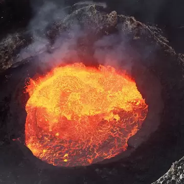 A circular lake of molten lava glowing orange inside a dark volcanic crater, steam rising from the rim.