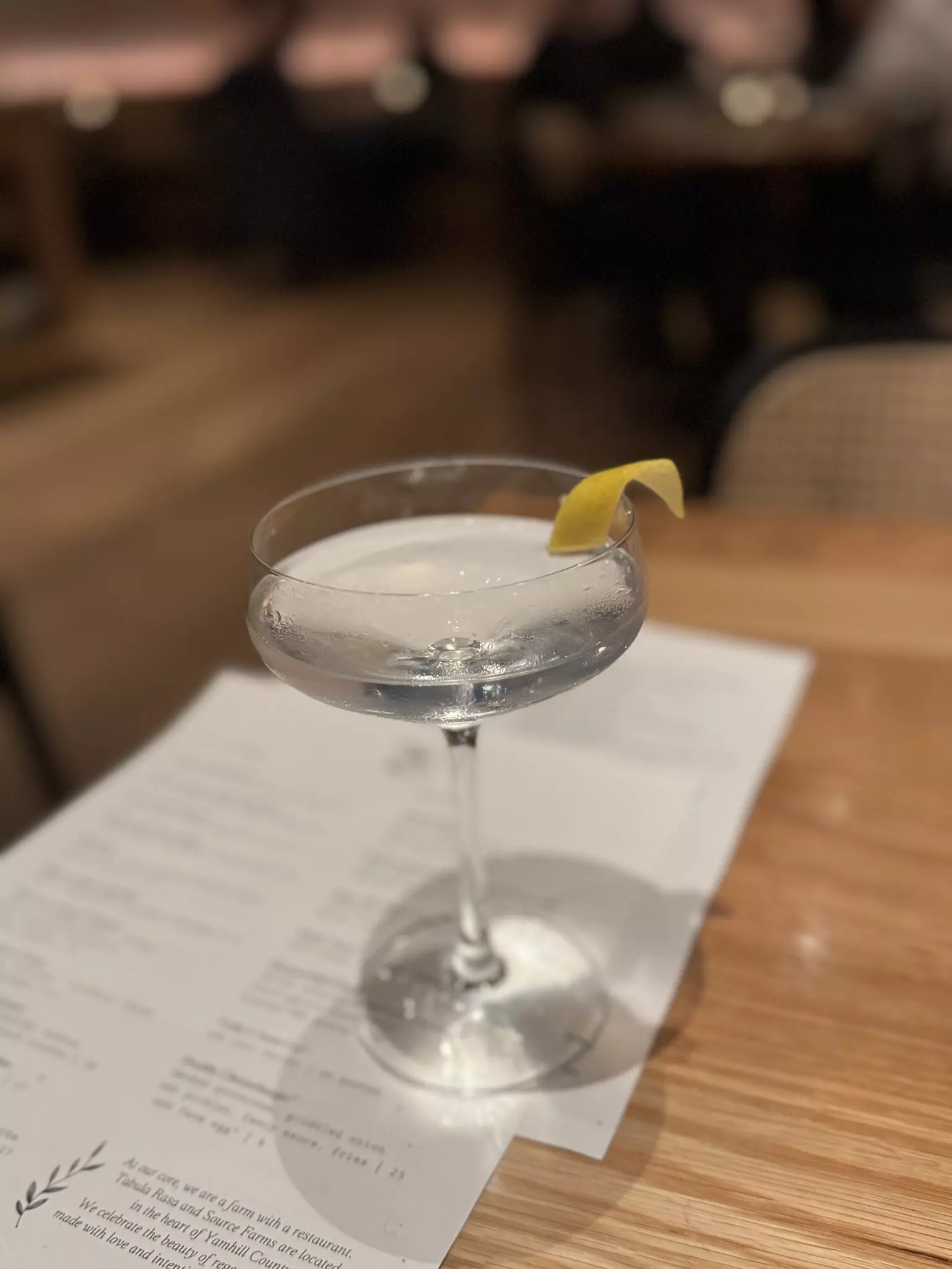 A martini glass filled with clear liquid and a sprig of lemon sitting atop a menu on a wooden table.