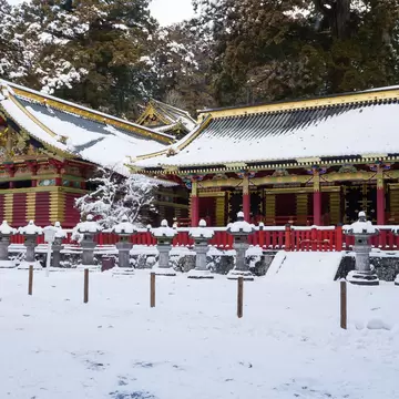 Red and gold shrine buildings with snowy roofs behind a row of snow-capped stone lanterns and cedar trees in Nikko.
