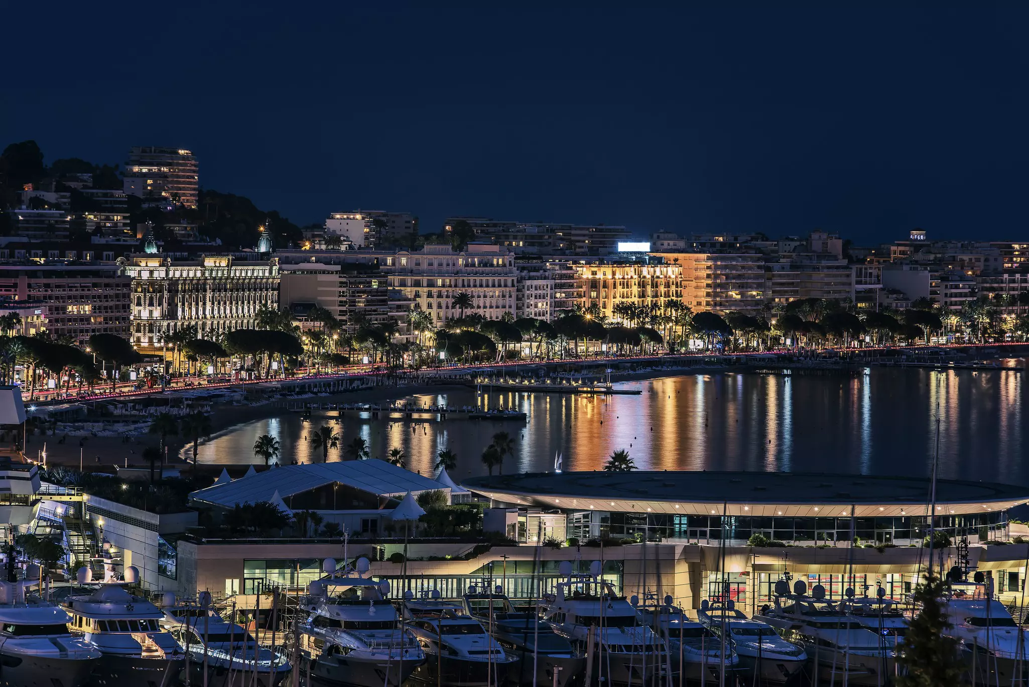 A wide view of a port city by night. Hotels and apartment buildings line the waterfront, their lights reflected in the still water opposite. Boats moored at a pier are visible in the front of the scene.