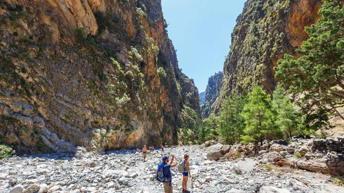 Hikers pause to take photos as they follow a rocky trail through a gorge between two vast cliff faces.