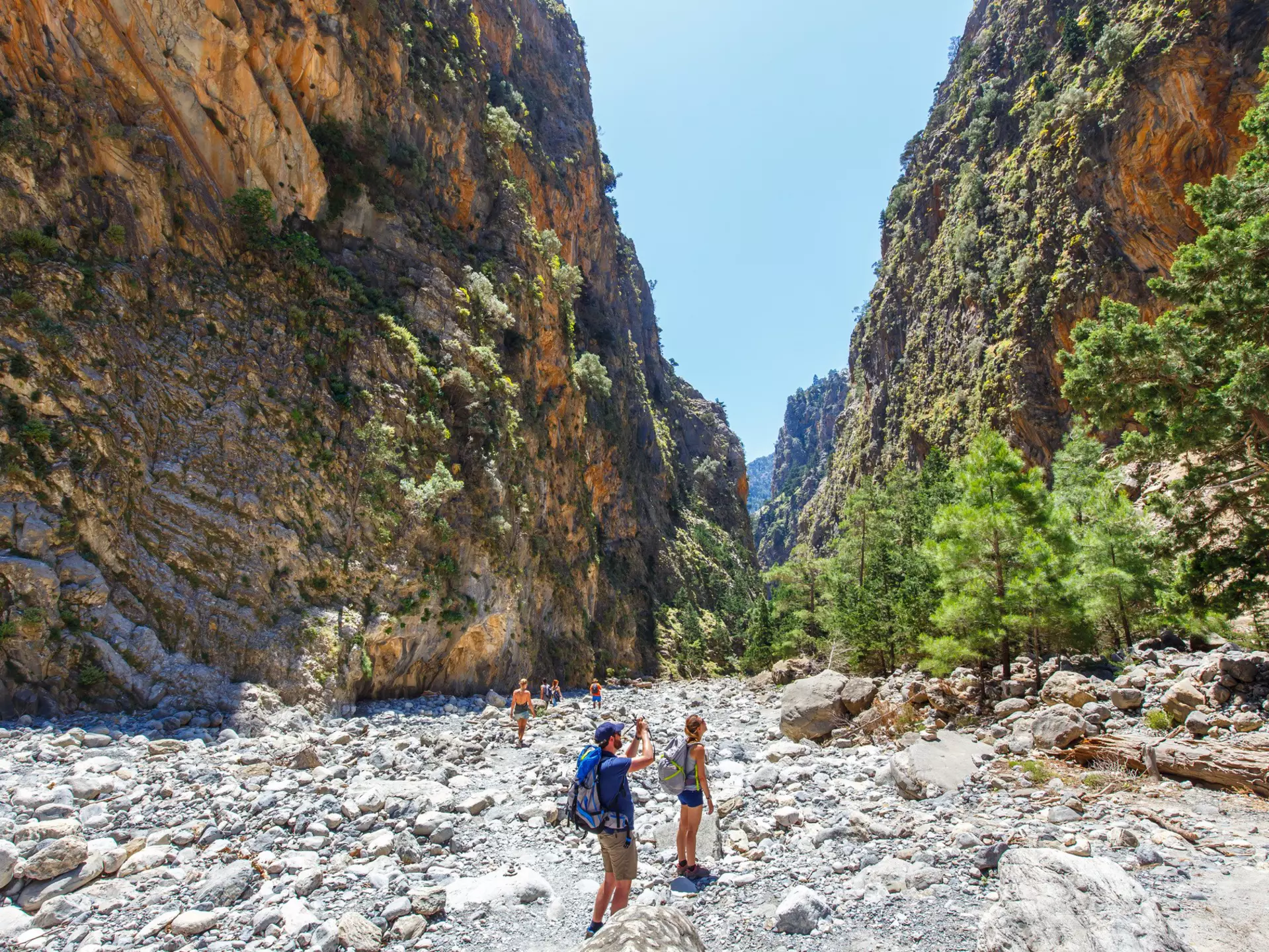 Hikers pause to take photos as they follow a rocky trail through a gorge between two vast cliff faces.