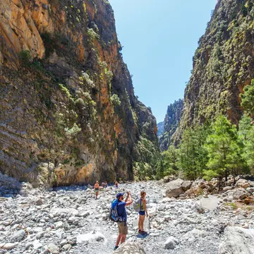 Hikers pause to take photos as they follow a rocky trail through a gorge between two vast cliff faces.