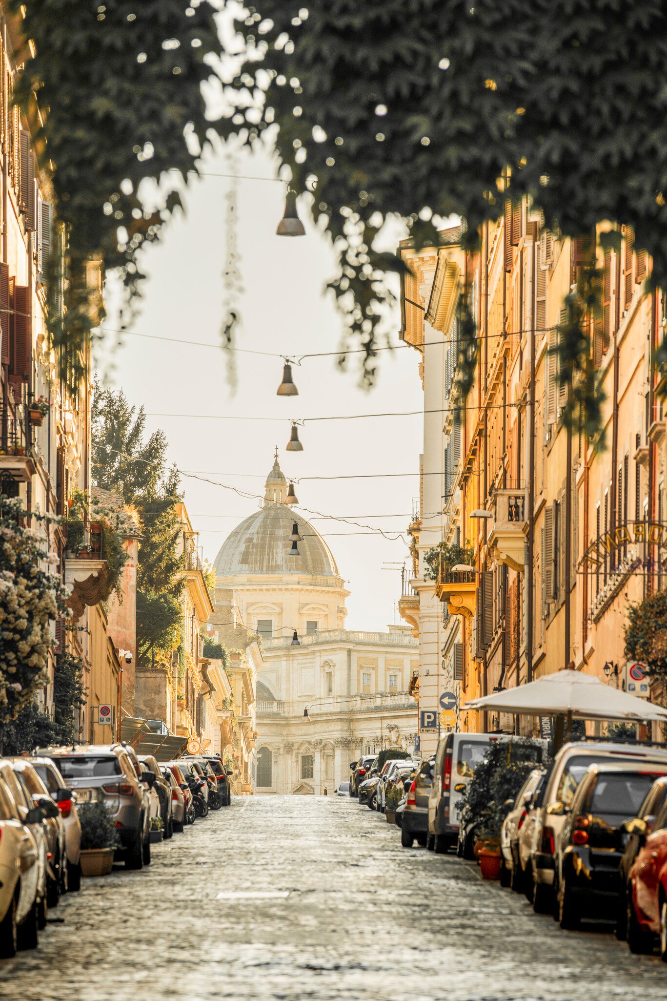 A cobbled street in Rome's Monti lined with parked cars and hung with lamps and ivy, leading to a church dome in hazy light.