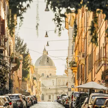 A cobbled street in Rome's Monti lined with parked cars and hung with lamps and ivy, leading to a church dome in hazy light.