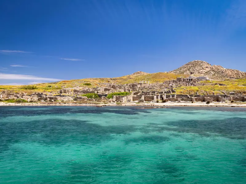 A view of the ancient ruins on the island of Delos with crystal clear blue water in the foreground. 