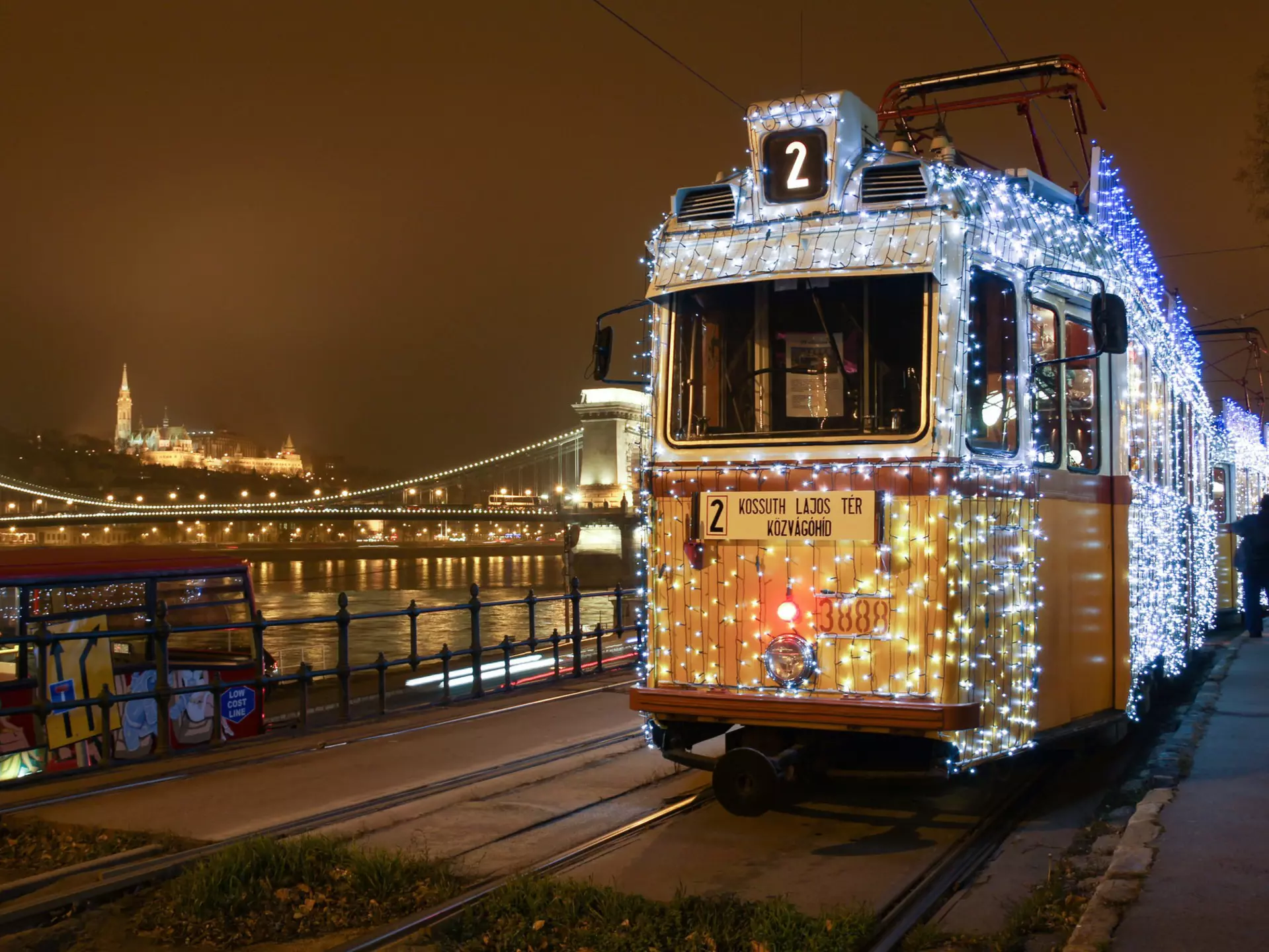 A shot from the front of a tram covered in small white and blue lights, stopped near the river