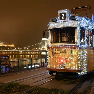 A shot from the front of a tram covered in small white and blue lights, stopped near the river
