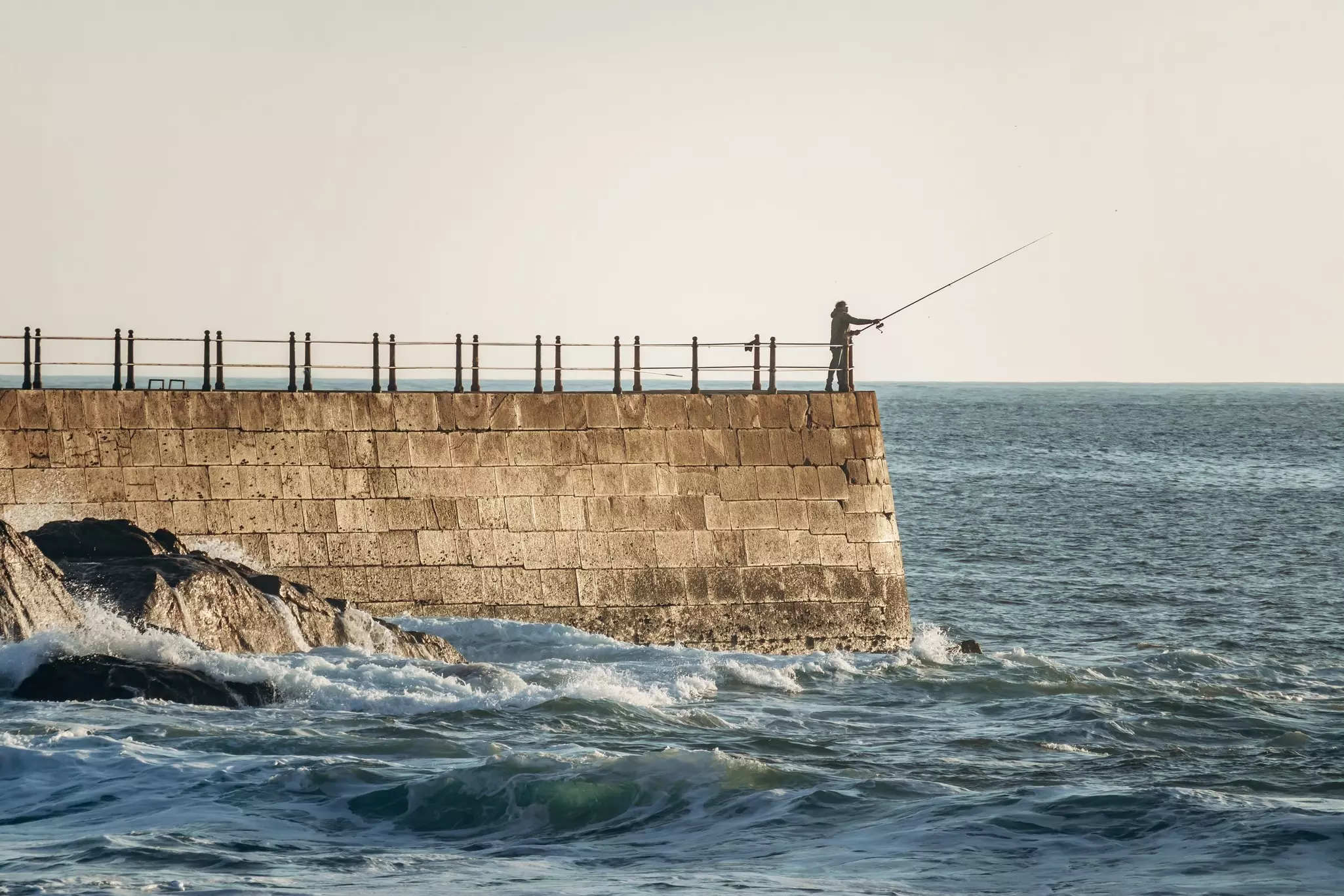 A fisherman fishing from a tall stone pier in the Atlantic Ocean with crashing waves in the foreground.