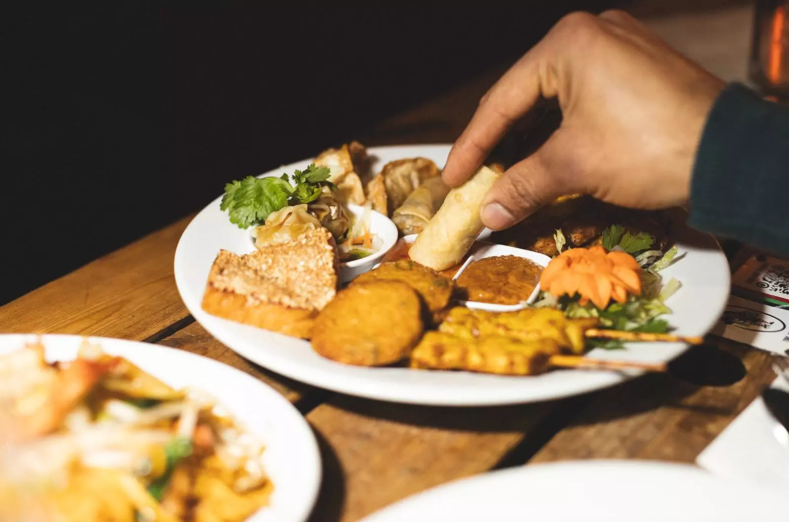 A hand reaches out for a spring roll on a plate of Thai appetizers.
