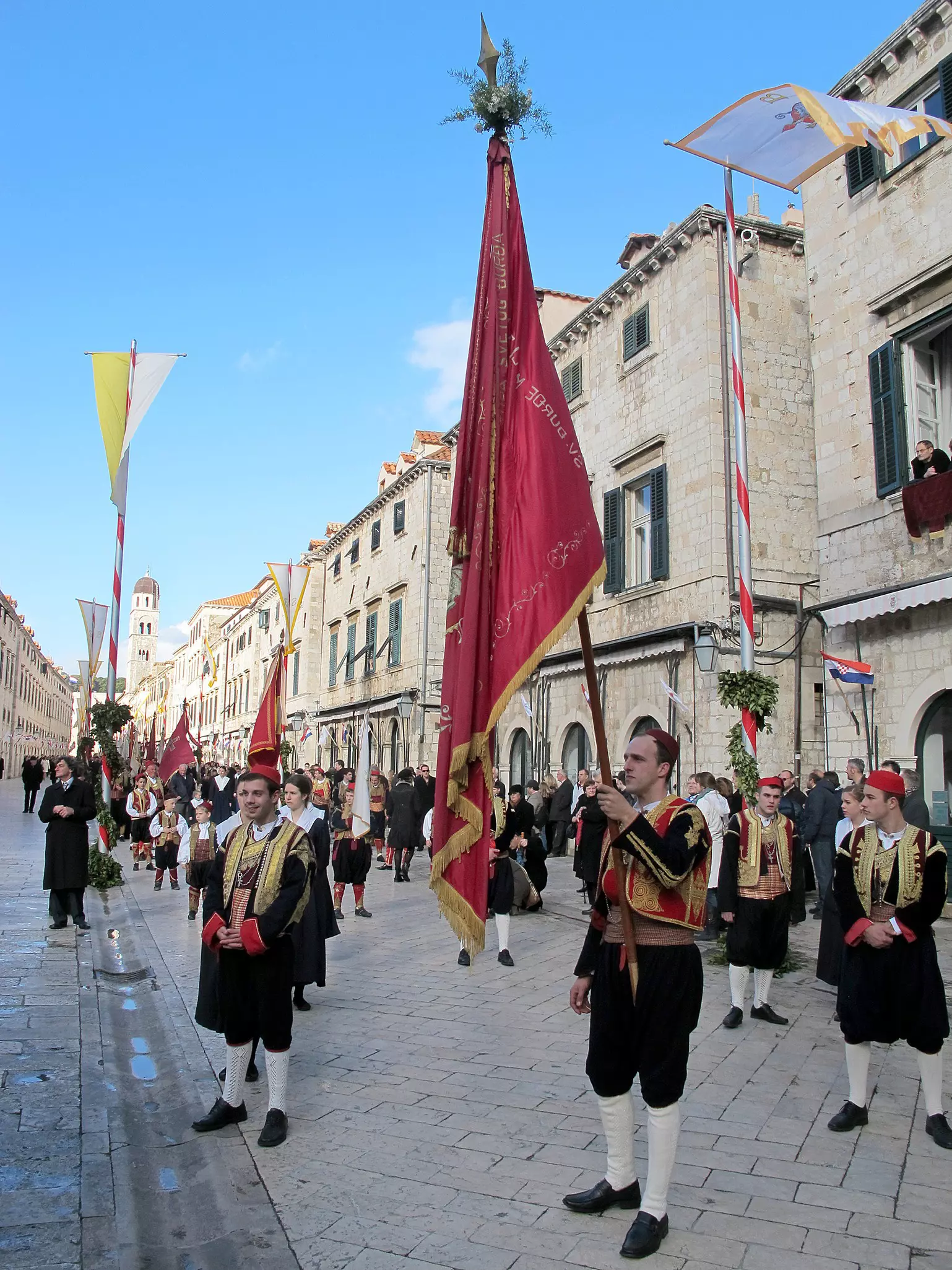 People in national dress stand in an old town holding flags as part of a festival parade.