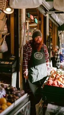  A vendor with a long red beard stands behind a fresh fruit stand 
