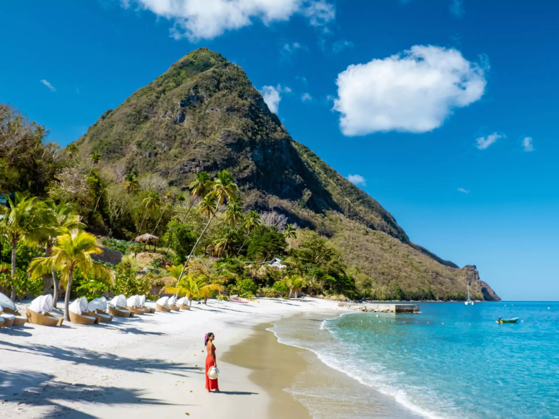 A woman in a full-length red sundress stands on beach backed by lush greenery with a tall pointed mountain at the end of the cove.