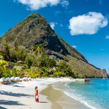 A woman in a full-length red sundress stands on beach backed by lush greenery with a tall pointed mountain at the end of the cove.