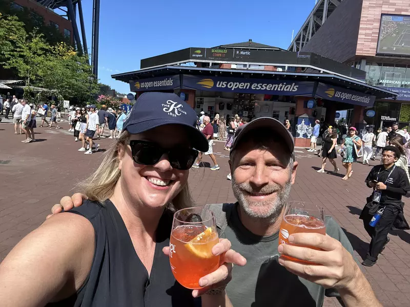 A woman and a man, both wearing baseball caps to shade their eyes from the sun, stand in front of a building with a sign that says "US Open Essentials" 