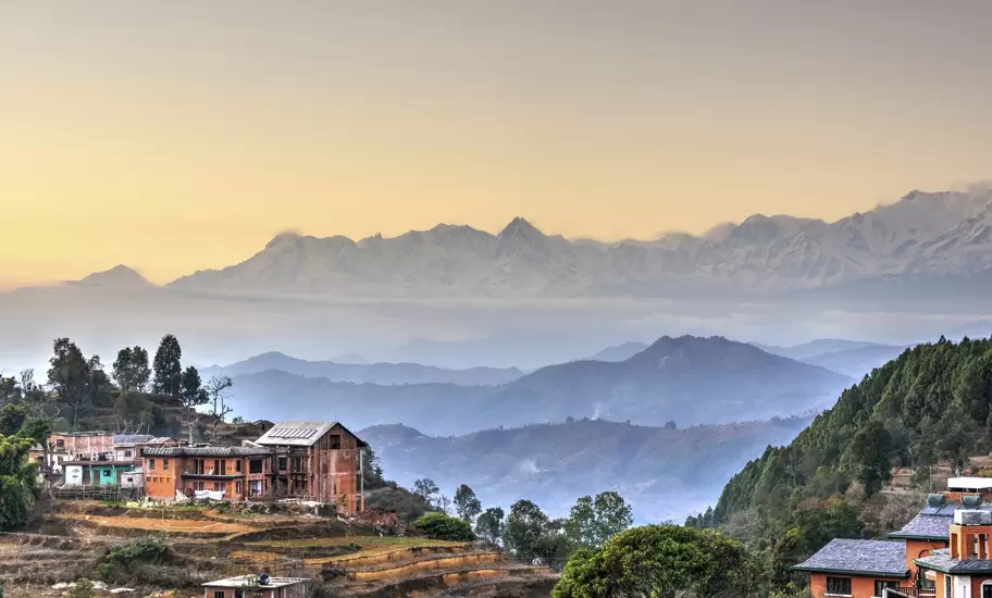 Brick houses on terraced hillside in Bandipur, Nepal, with layered blue ridges and a snowcapped Himalayan range at sunrise