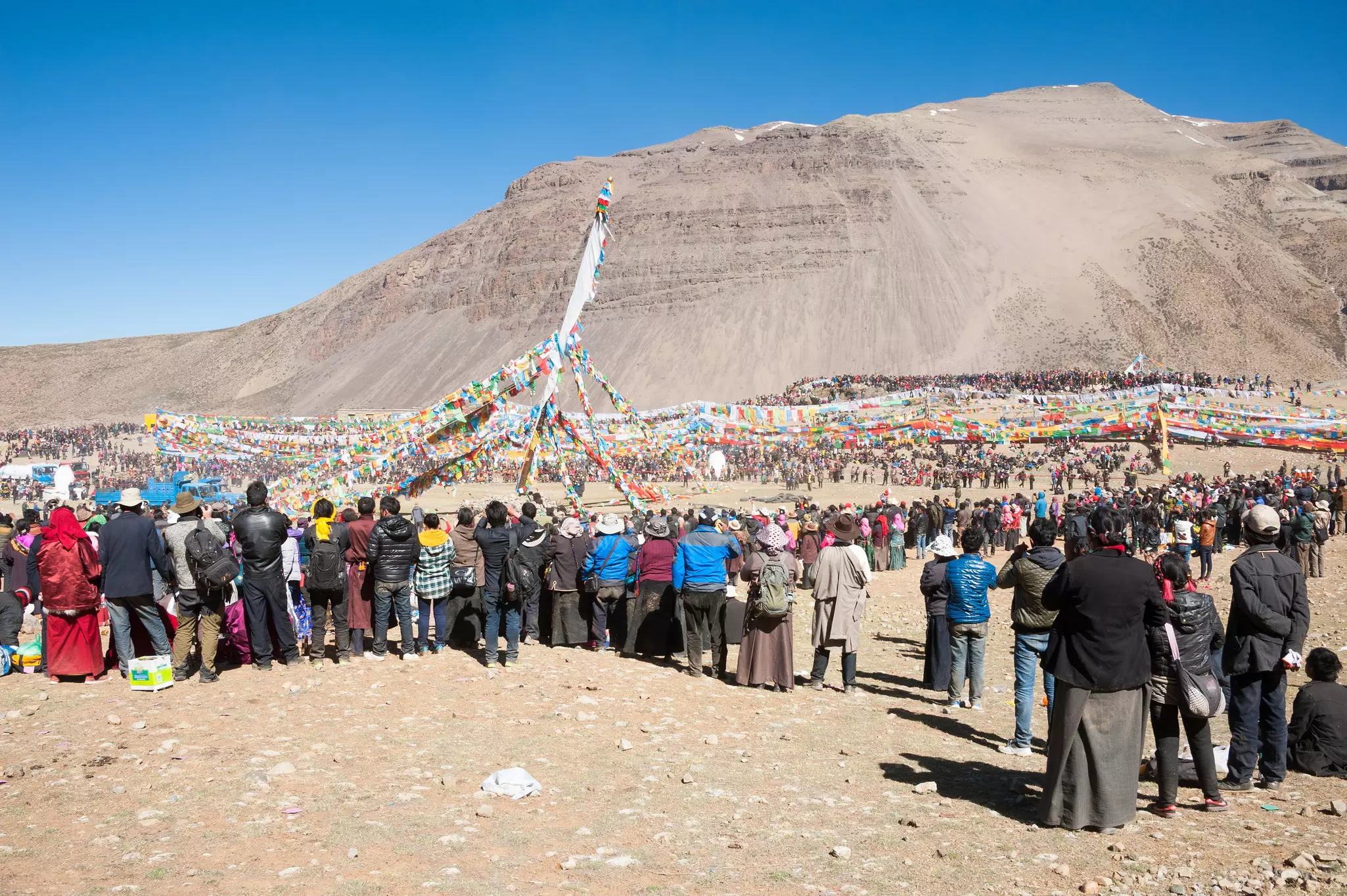 Crowds of people gather to watch a festival held on a mountain plateau under colorful prayer flags.
