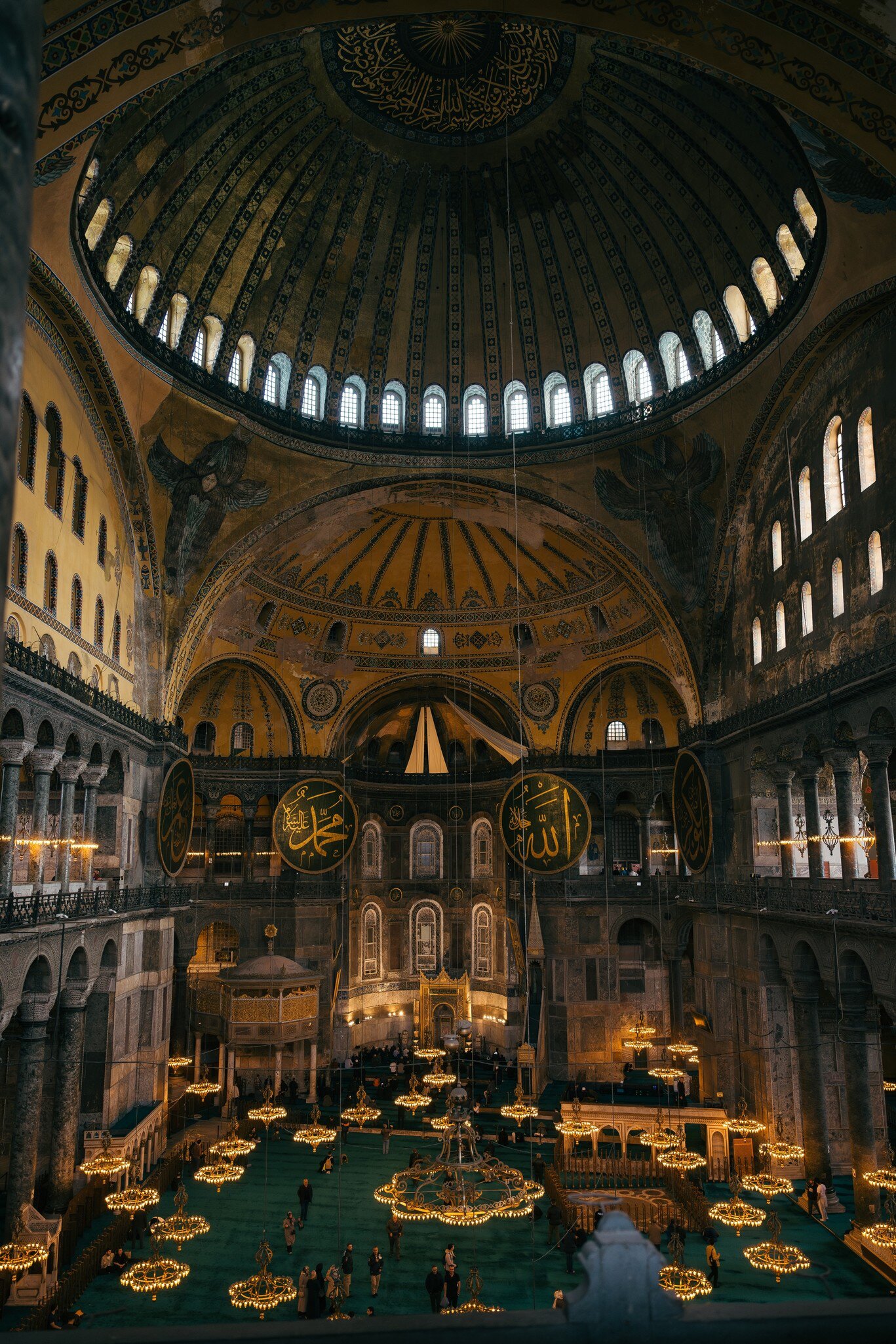 Ribbed dome on a ring of windows above seraph mosaics, gold calligraphic roundels and low chandeliers over green carpet.