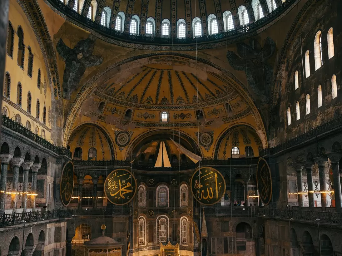 Ribbed dome on a ring of windows above seraph mosaics, gold calligraphic roundels and low chandeliers over green carpet.